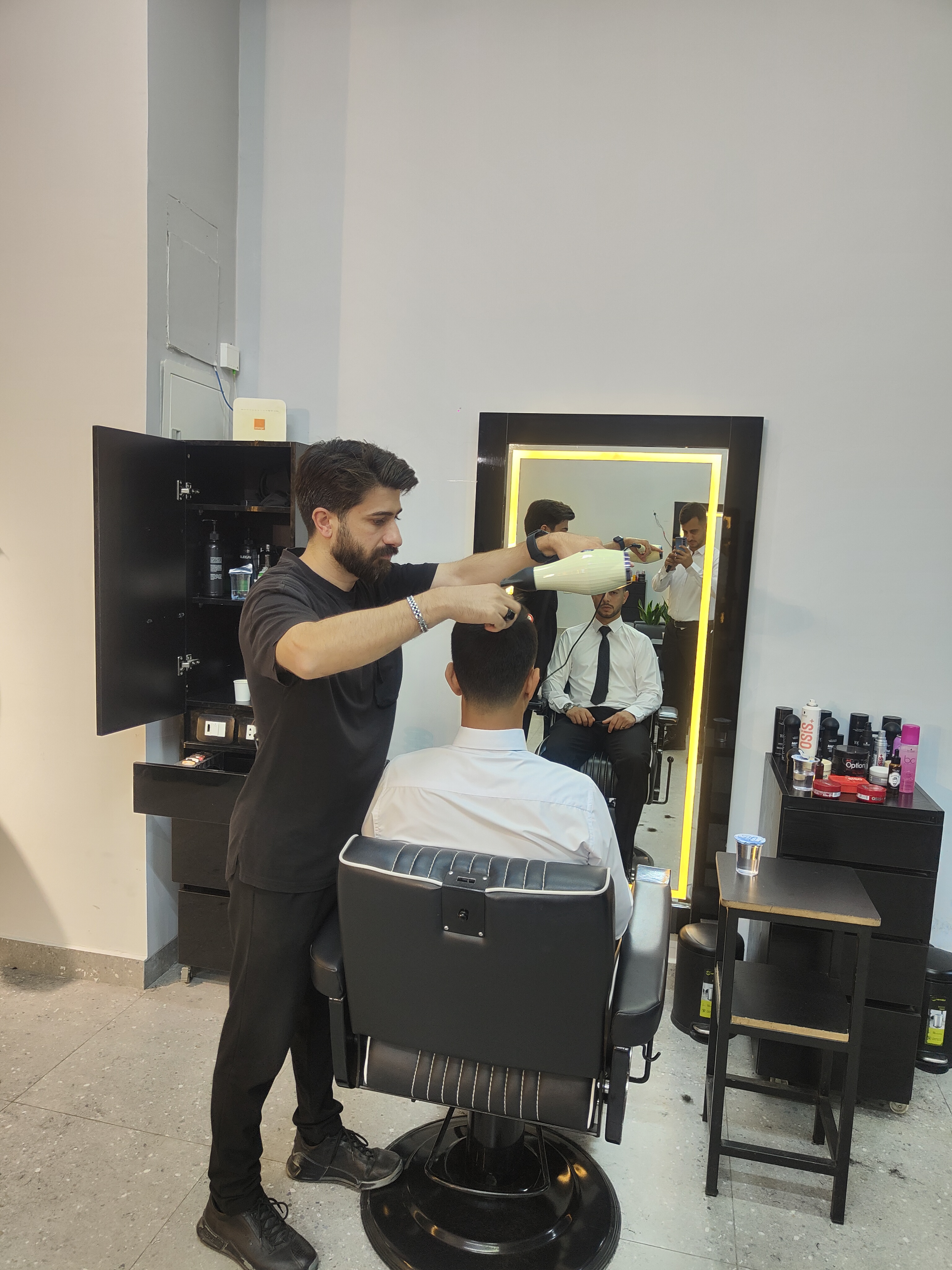 Barber blow-drying a client's hair beside a backlit gold-framed mirror